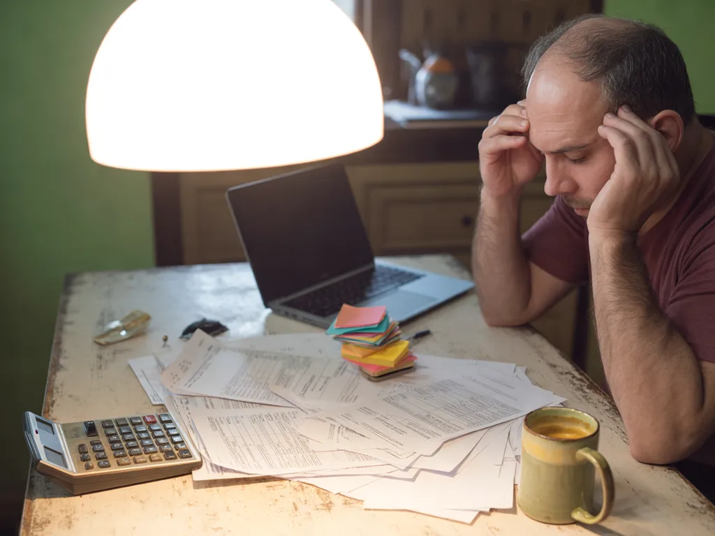 A tired owner with his head in his hands at a kitchen table covered in invoices, sticky notes and a calculator, late at night